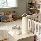 A cream-colored vacuum insulated bottle with a carrying loop sits on a white bedside table next to two glass baby bottles filled with milk in a softly lit nursery room.